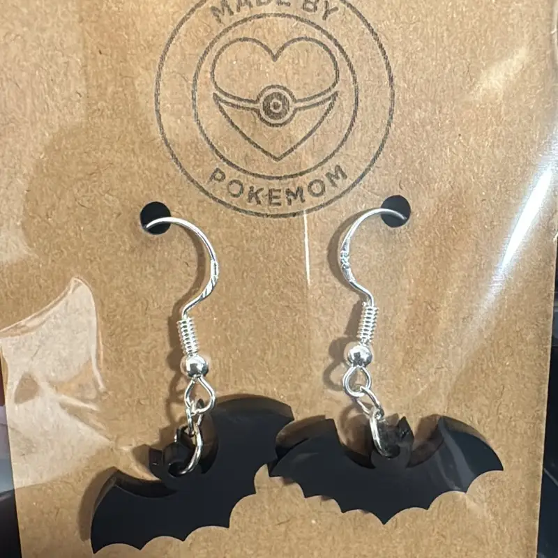 A pair of matte black acrylic bat earrings on silver-tone hooks, displayed on a white card inside a glass bell jar.