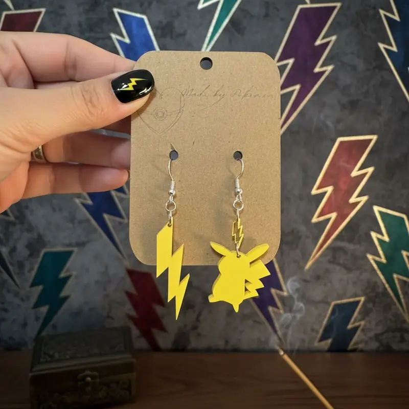 Mismatched acrylic earrings on a kraft card: a yellow Pikachu-inspired head and a separate lightning bolt, held against a desk.