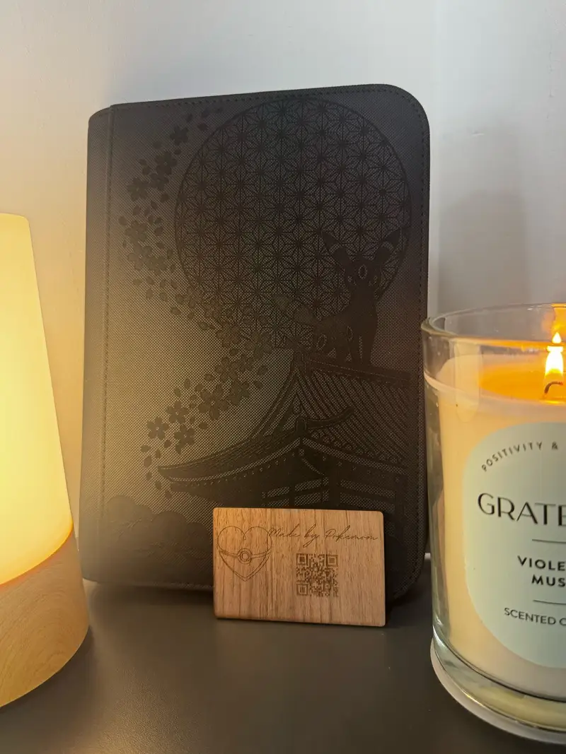 Black binder engraved in gold with an Umbreon-inspired figure on a temple roof, a geometric moon, falling blossom, and the title The Temple Watch. A lit candle and a small card sit beside the binder.